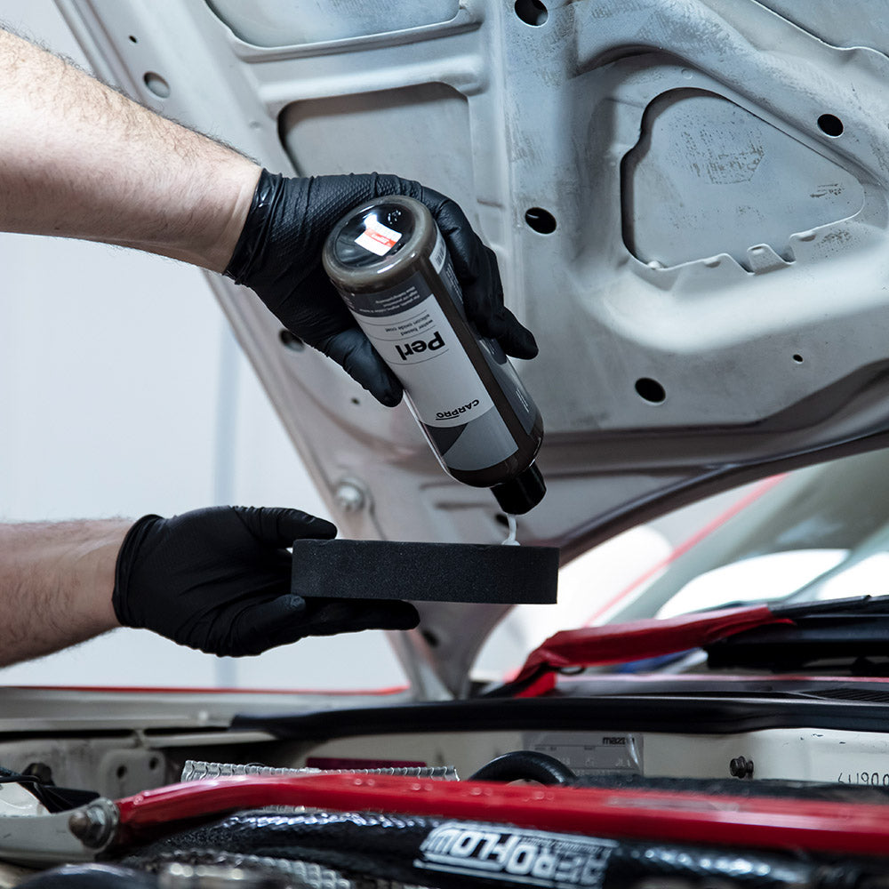 Gloved hands pouring CARPRO PERL onto a foam applicator in an engine bay