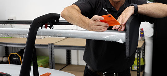 A person using an orange PPF squeege to apply PPF to a spoiler